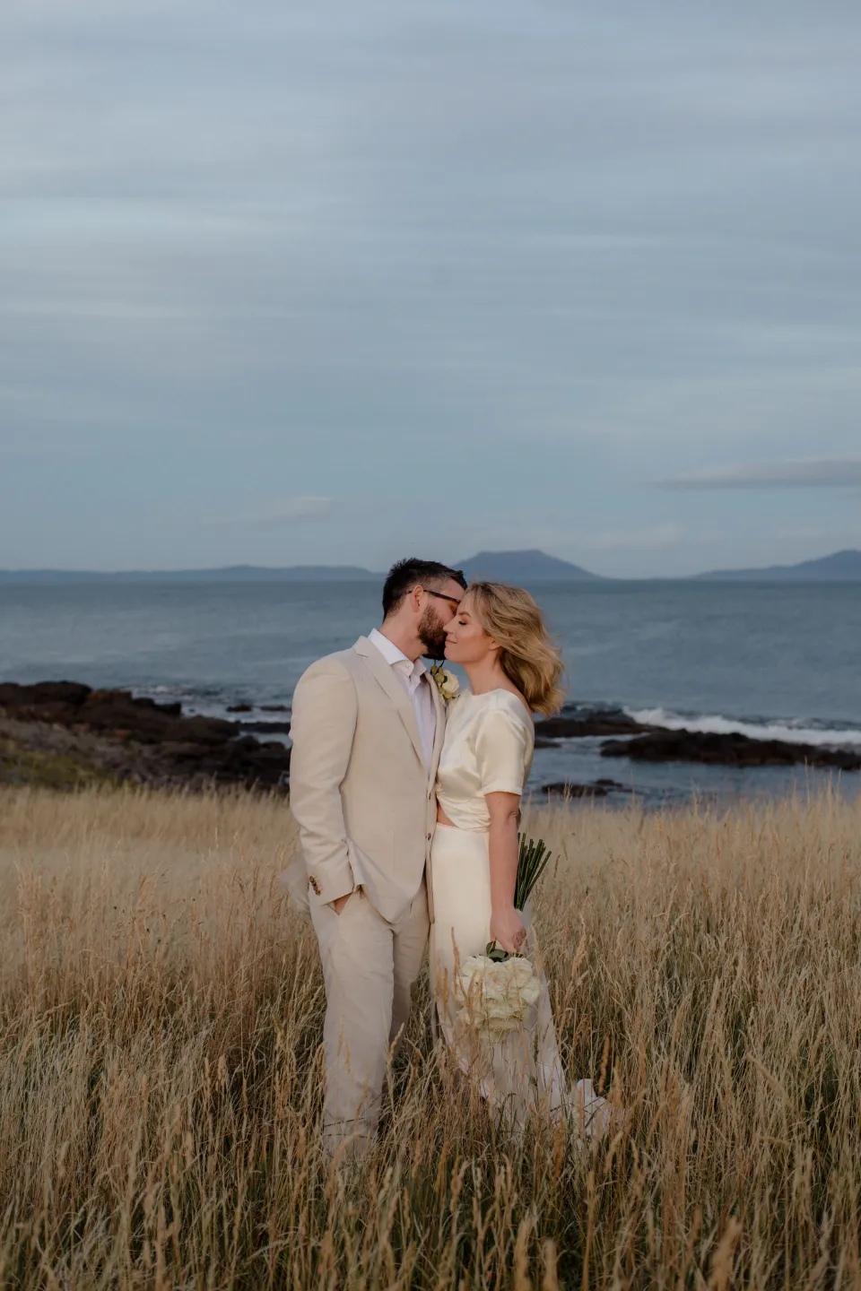 Jemmah and Jack kissing among the golden grasses, at Thalia Haven on the East Coast of Tasmania, Australia, for an Elopement Collective elopement. Photograph by Joey Willis.