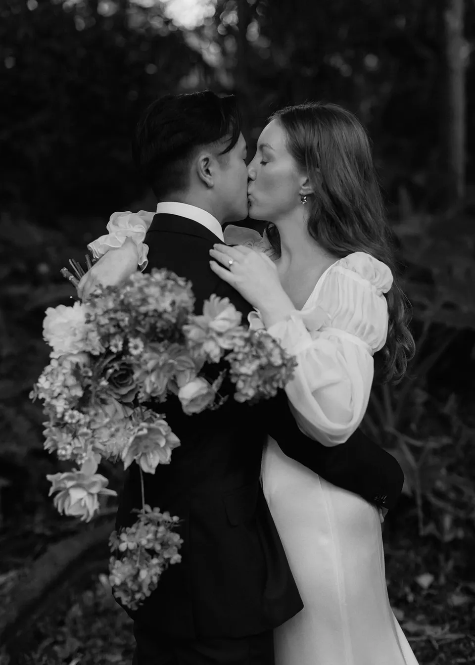 Black-and-white photograph of the couple kissing with the bouquet between them, with flowers by The Perfect Party Co. At Fig Tree Villa in Ewingsdale, Byron Bay, for an Elopement Collective elopement. Photograph by Bulb Creative.