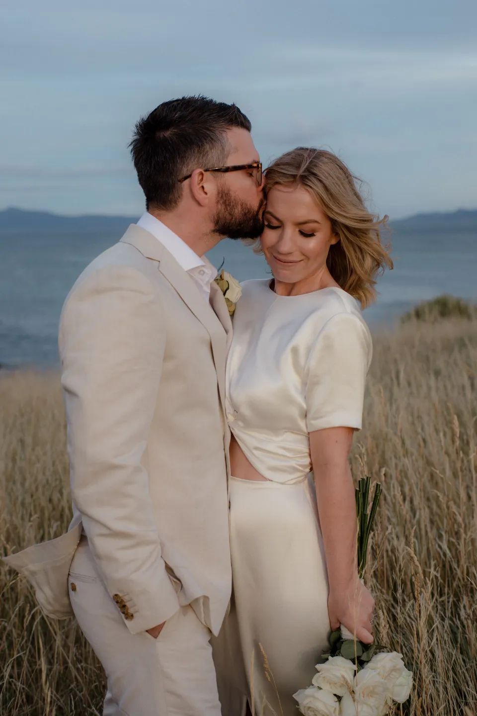 Jack kissing Jemmah’s forehead with the blue bay behind them, at Thalia Haven on the East Coast of Tasmania, Australia, for an Elopement Collective elopement. Photograph by Joey Willis.