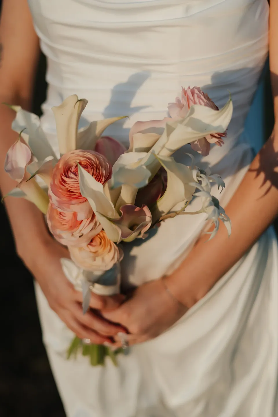 Close-up of Dani holding her peach and cream bouquet by The Romantics against the folds of her white dress, at Thalia Haven on Tasmania’s east coast, for an Elopement Collective elopement. Photograph by Bulb Creative.