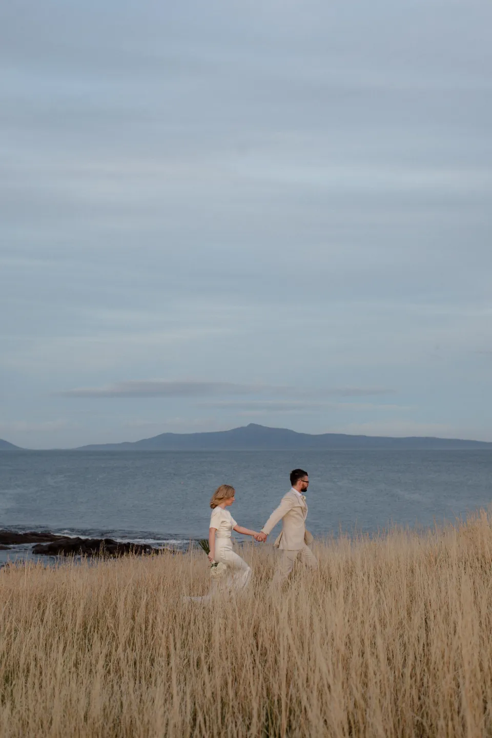 A wide view of Jemmah and Jack standing together in tall grass above the coast, at Thalia Haven on the East Coast of Tasmania, Australia, for an Elopement Collective elopement. Photograph by Joey Willis.