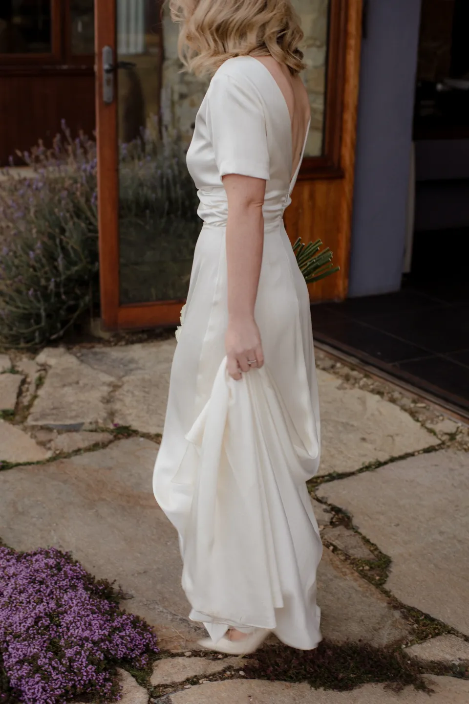 A close view of Jemmah’s dress as she stands among stone paving and purple garden flowers, at Thalia Haven on the East Coast of Tasmania, Australia, for an Elopement Collective elopement. Photograph by Joey Willis.