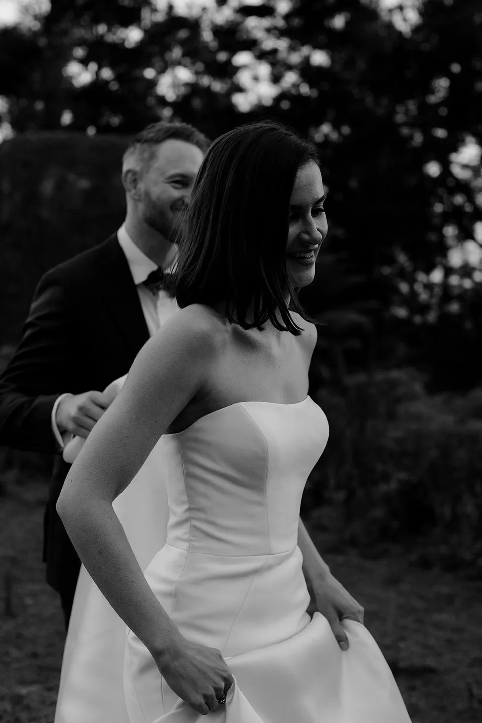 Black and white portrait of Meg walking ahead of Ryan beneath the trees. At The Keep on Tasmania’s east coast, for an Elopement Collective elopement. Photograph by Jenna Agius.