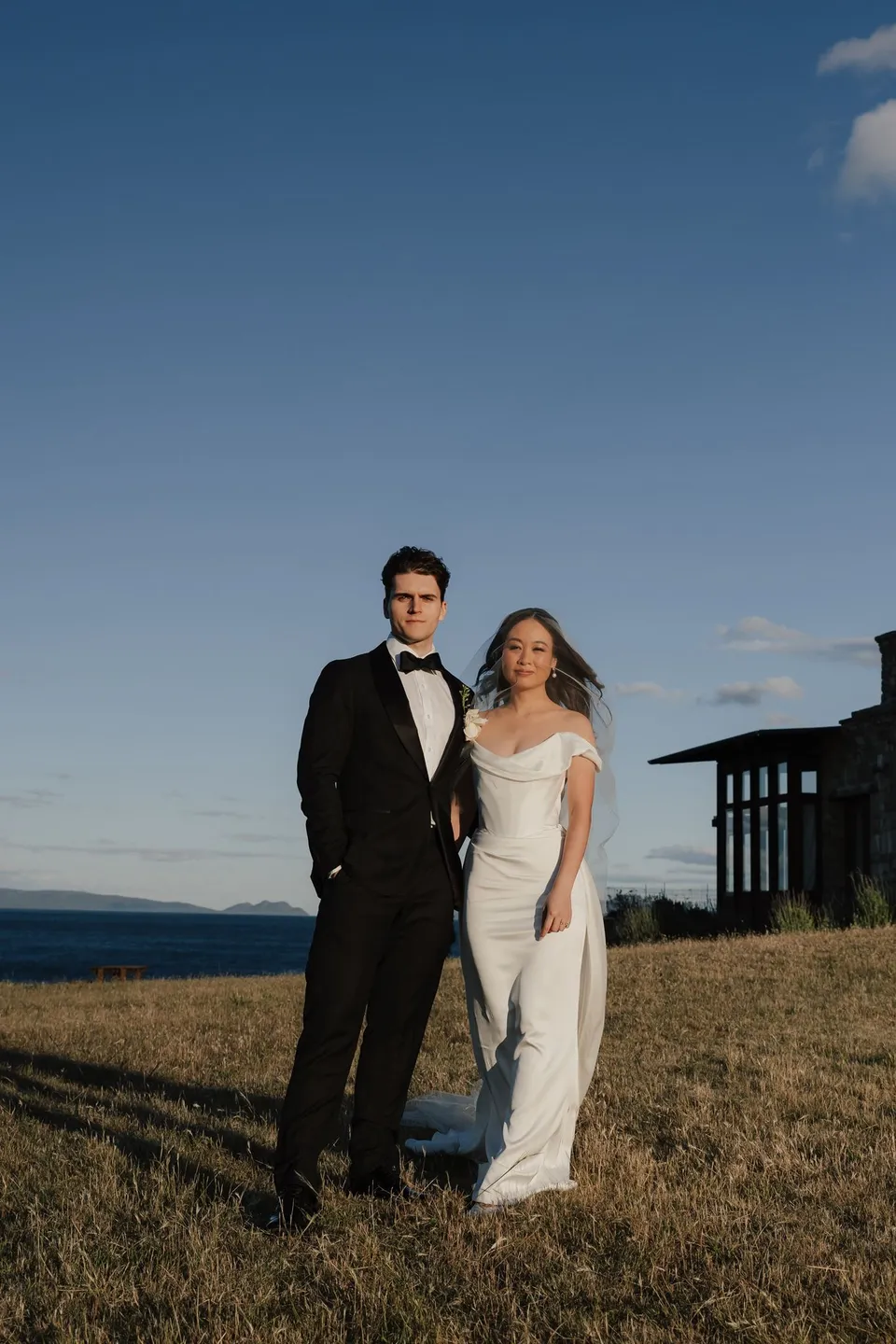Clara and Harry standing together in golden grassland with the blue bay and stone house behind them, at Thalia Haven on the East Coast of Tasmania, for an Elopement Collective elopement. Photograph by Bulb Creative.