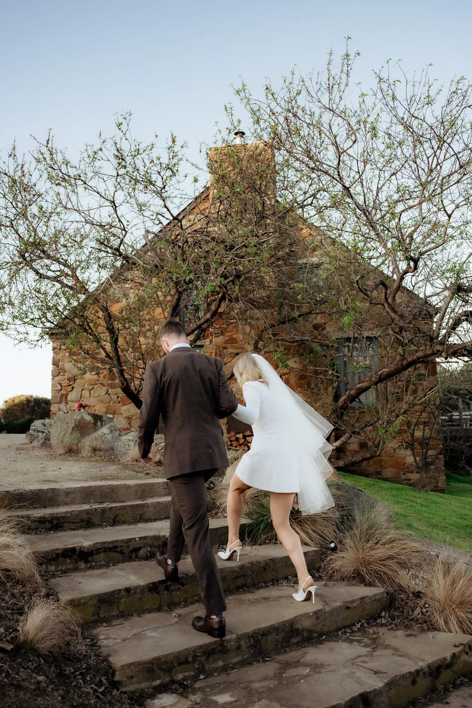 Rear view of Kaitlyn and Russell climbing stone steps towards their cottage, with her veil blowing behind her. At Piermont Retreat on Tasmania’s east coast, for an Elopement Collective elopement. Photograph by Omar of By The Wilde.