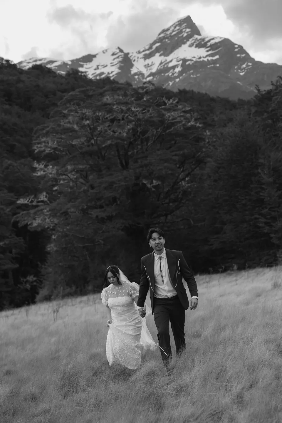 Jaeda and Adrian laughing and running hand in hand through the meadow in black and white, in the field on the road to Paradise near Glenorchy, in the Queenstown region of New Zealand, for an Elopement Collective Queenstown elopement. Photograph by Bulb Creative.