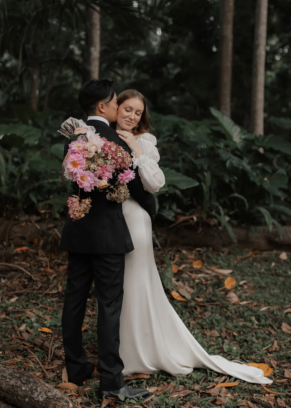 Elyse smiling towards the camera while embracing Jonathan beneath the trees. At Fig Tree Villa in Ewingsdale, Byron Bay, for an Elopement Collective elopement. Photograph by Bulb Creative.