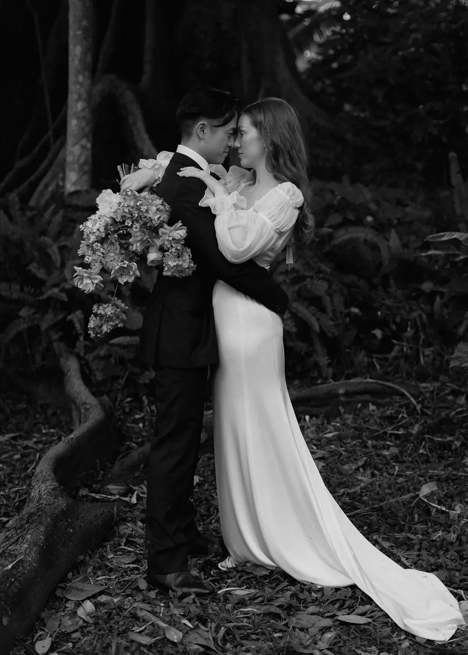 Black-and-white full-length portrait of the couple embracing beside a tree. At Fig Tree Villa in Ewingsdale, Byron Bay, for an Elopement Collective elopement. Photograph by Bulb Creative.