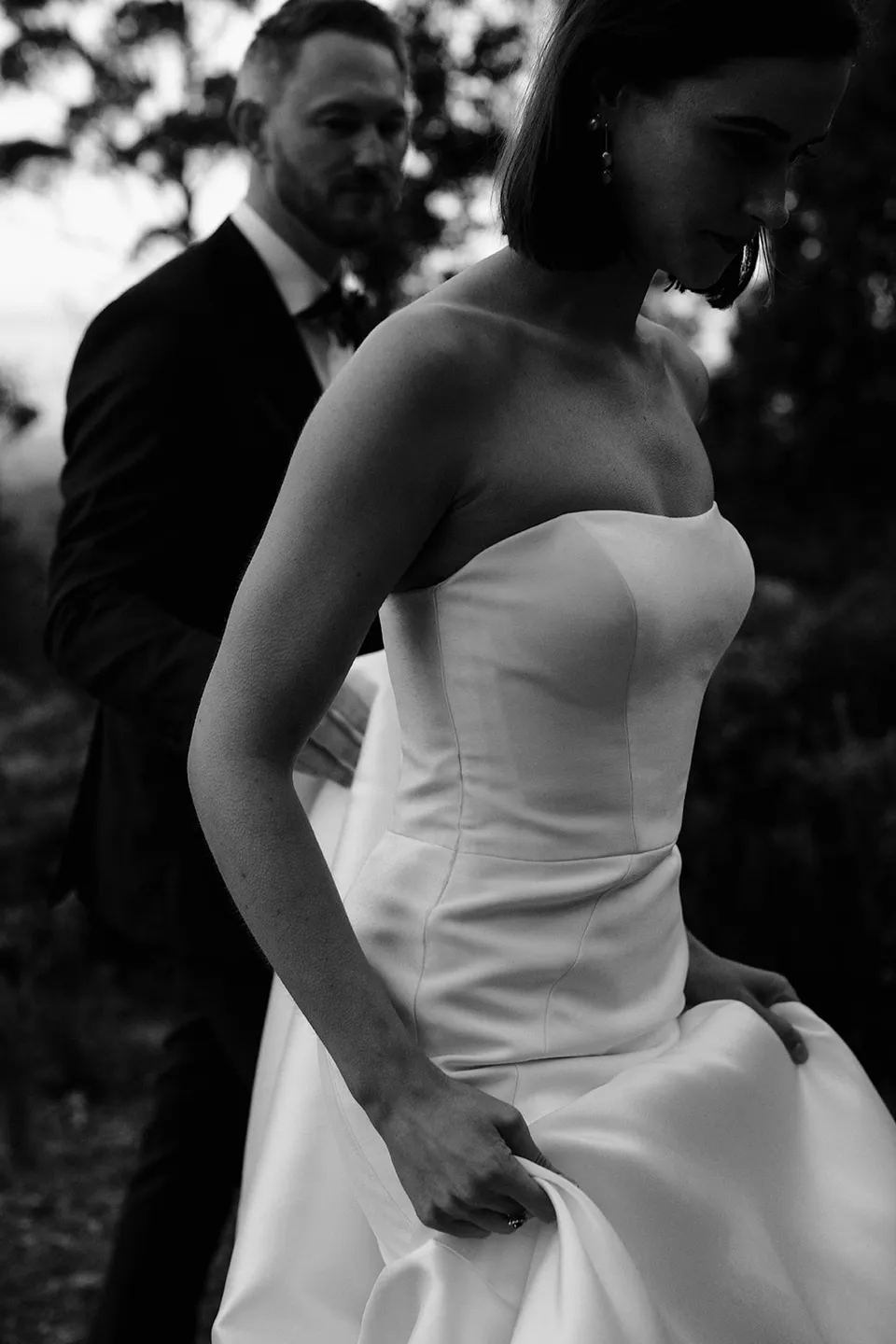 Black and white close view of Meg gathering her wedding dress as Ryan follows through the woodland. At The Keep on Tasmania’s east coast, for an Elopement Collective elopement. Photograph by Jenna Agius.