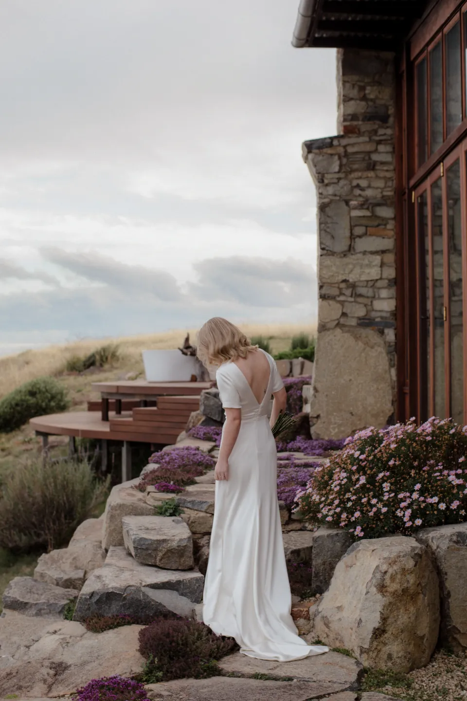 Jemmah with her bouquet by The Romantics beside the stone house and outdoor bath, at Thalia Haven on the East Coast of Tasmania, Australia, for an Elopement Collective elopement. Photograph by Joey Willis.