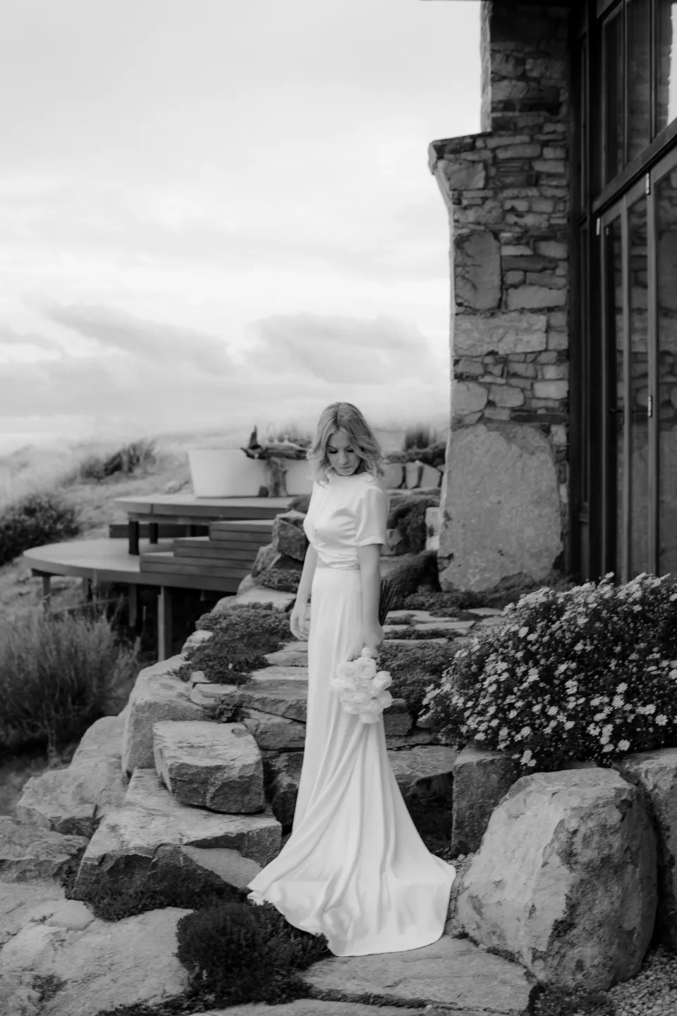 Black and white portrait of Jemmah standing on stone steps beside the house, at Thalia Haven on the East Coast of Tasmania, Australia, for an Elopement Collective elopement. Photograph by Joey Willis.