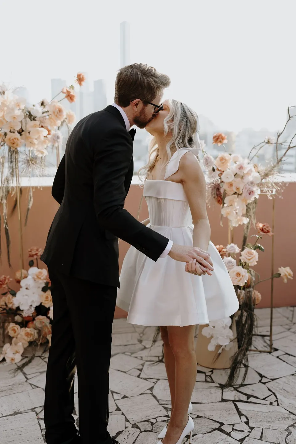 Danielle and Adam kissing on the terrace with peach and white flowers behind them, during Danielle and Adam’s Elopement Collective elopement at The Calile Hotel in Fortitude Valley, Brisbane.