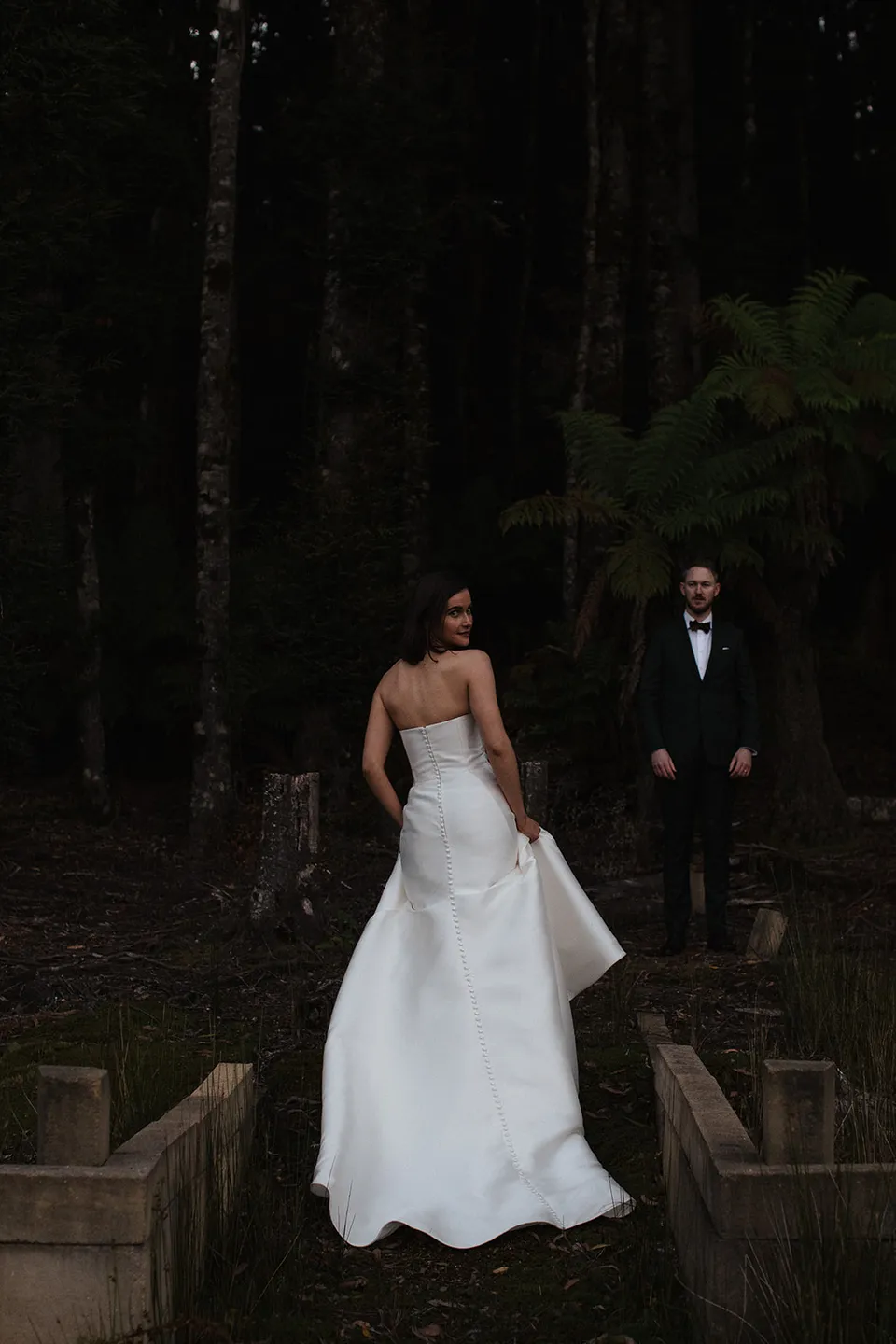 Meg stepping across a small bridge with Ryan behind her, surrounded by dark forest and ferns. At The Keep on Tasmania’s east coast, for an Elopement Collective elopement. Photograph by Jenna Agius.