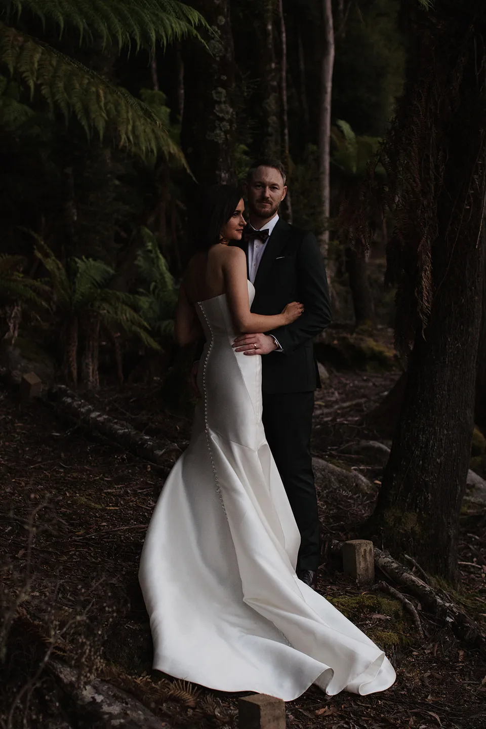 Meg and Ryan embracing among tall tree trunks and ferns, with her white dress falling to the forest floor. At The Keep on Tasmania’s east coast, for an Elopement Collective elopement. Photograph by Jenna Agius.