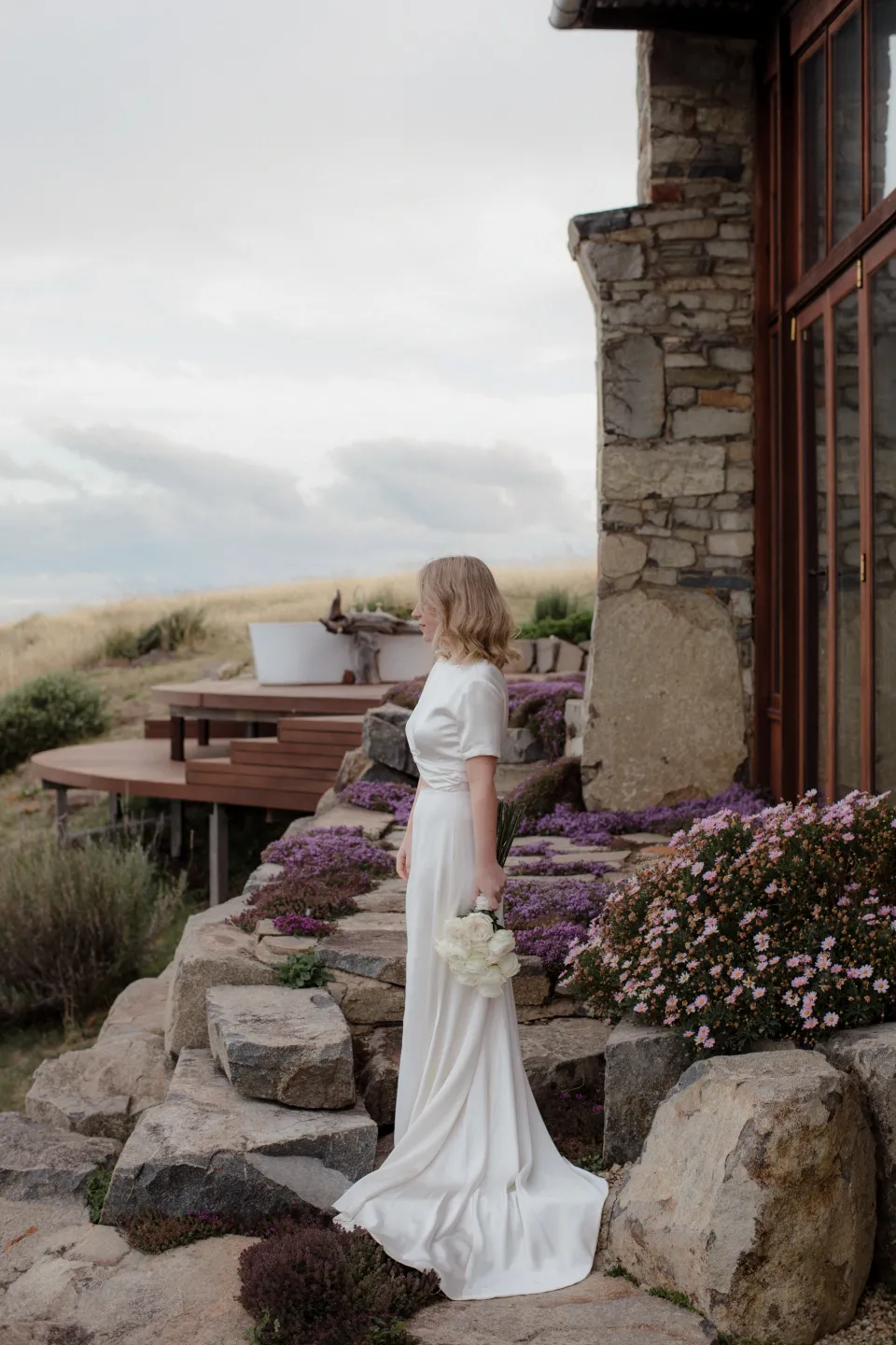 Jemmah looking towards the bay from the stone terrace, at Thalia Haven on the East Coast of Tasmania, Australia, for an Elopement Collective elopement. Photograph by Joey Willis.