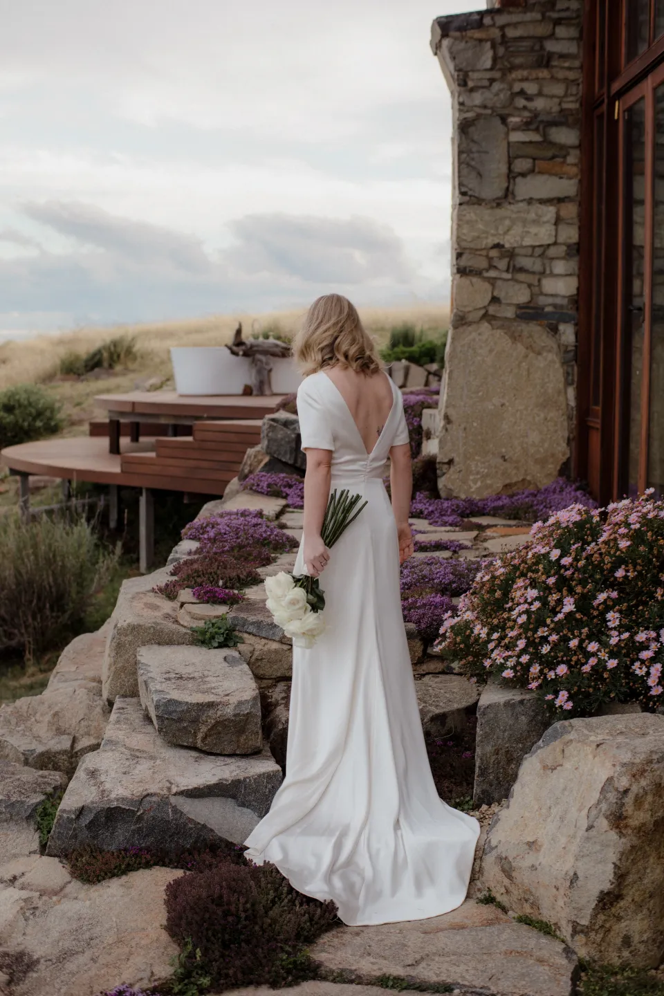 Jemmah holding her bouquet by The Romantics beside purple garden flowers, at Thalia Haven on the East Coast of Tasmania, Australia, for an Elopement Collective elopement. Photograph by Joey Willis.