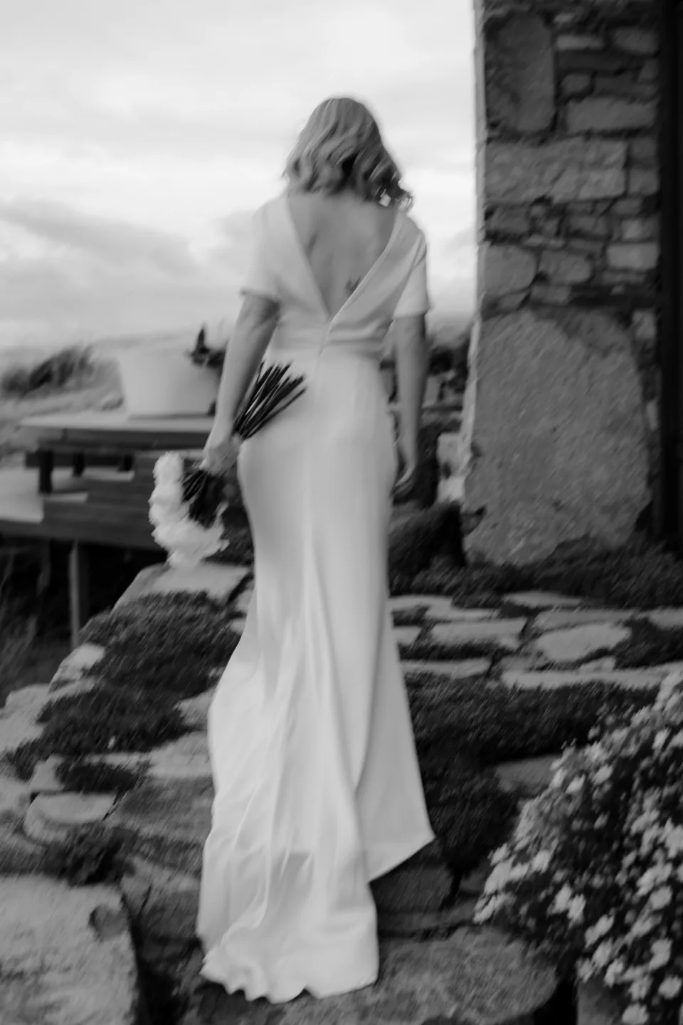 Black and white rear view of Jemmah beside the stone house, at Thalia Haven on the East Coast of Tasmania, Australia, for an Elopement Collective elopement. Photograph by Joey Willis.