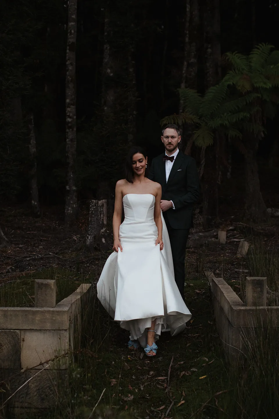 Meg and Ryan walking together across the small woodland bridge beneath tall trees. At The Keep on Tasmania’s east coast, for an Elopement Collective elopement. Photograph by Jenna Agius.