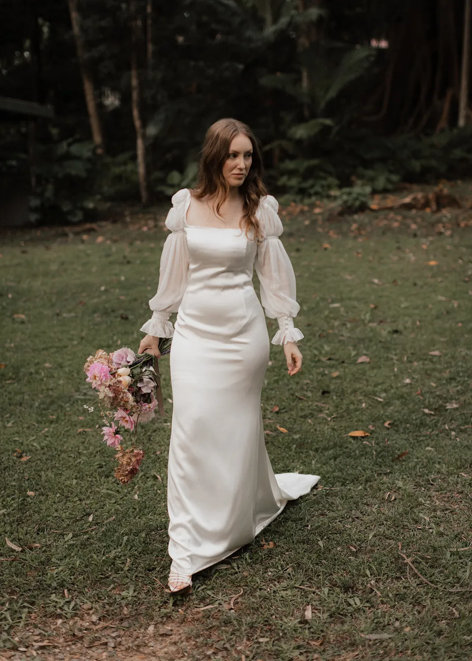 Elyse smiling while walking onto the lawn with flowers at her side. At Fig Tree Villa in Ewingsdale, Byron Bay, for an Elopement Collective elopement. Photograph by Bulb Creative.