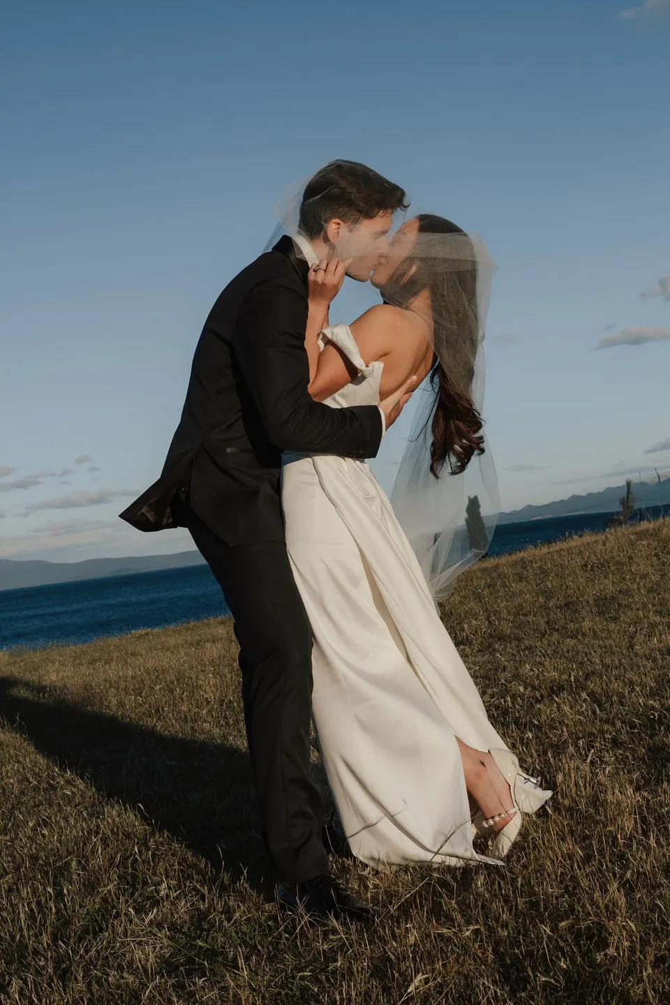 Harry lifting Clara into a kiss above the coastal grass, with her white dress moving around them, at Thalia Haven on the East Coast of Tasmania, for an Elopement Collective elopement. Photograph by Bulb Creative.