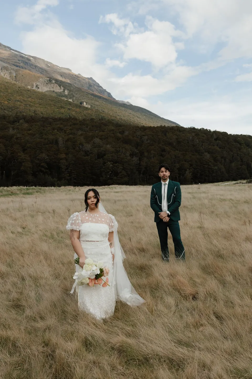 Jaeda holding her pink and white Orange Blossom Designs bouquet in the meadow, with Adrian and the mountains behind her, in the field on the road to Paradise near Glenorchy, in the Queenstown region of New Zealand, for an Elopement Collective Queenstown elopement. Photograph by Bulb Creative.