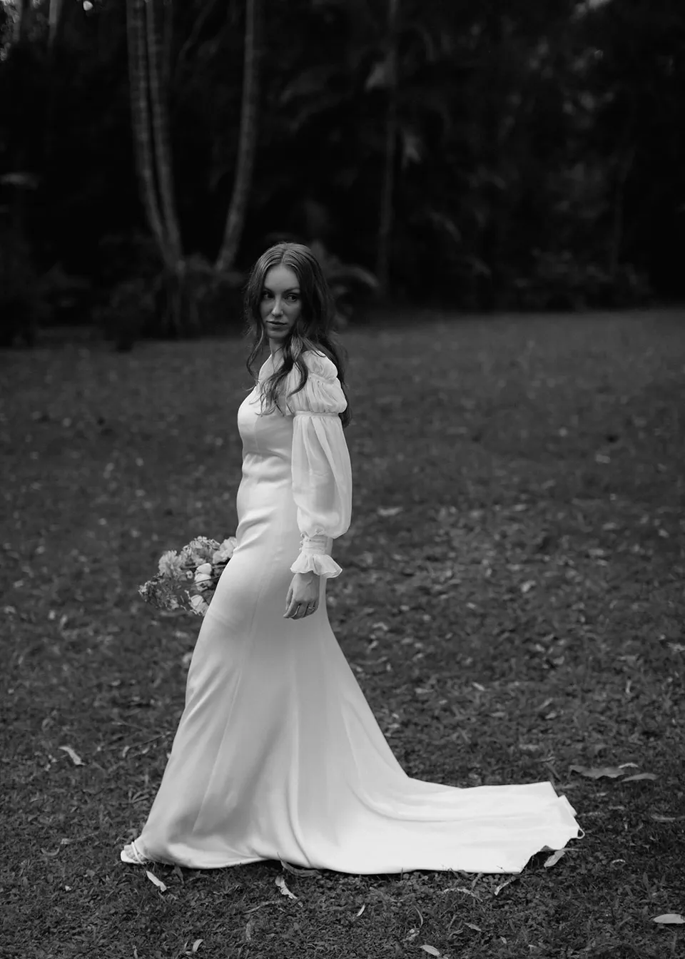 Black-and-white portrait of Elyse looking back across the lawn. At Fig Tree Villa in Ewingsdale, Byron Bay, for an Elopement Collective elopement. Photograph by Bulb Creative.