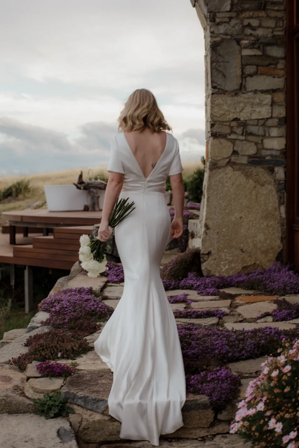 Jemmah standing beside the house with her dress train over the paving, at Thalia Haven on the East Coast of Tasmania, Australia, for an Elopement Collective elopement. Photograph by Joey Willis.