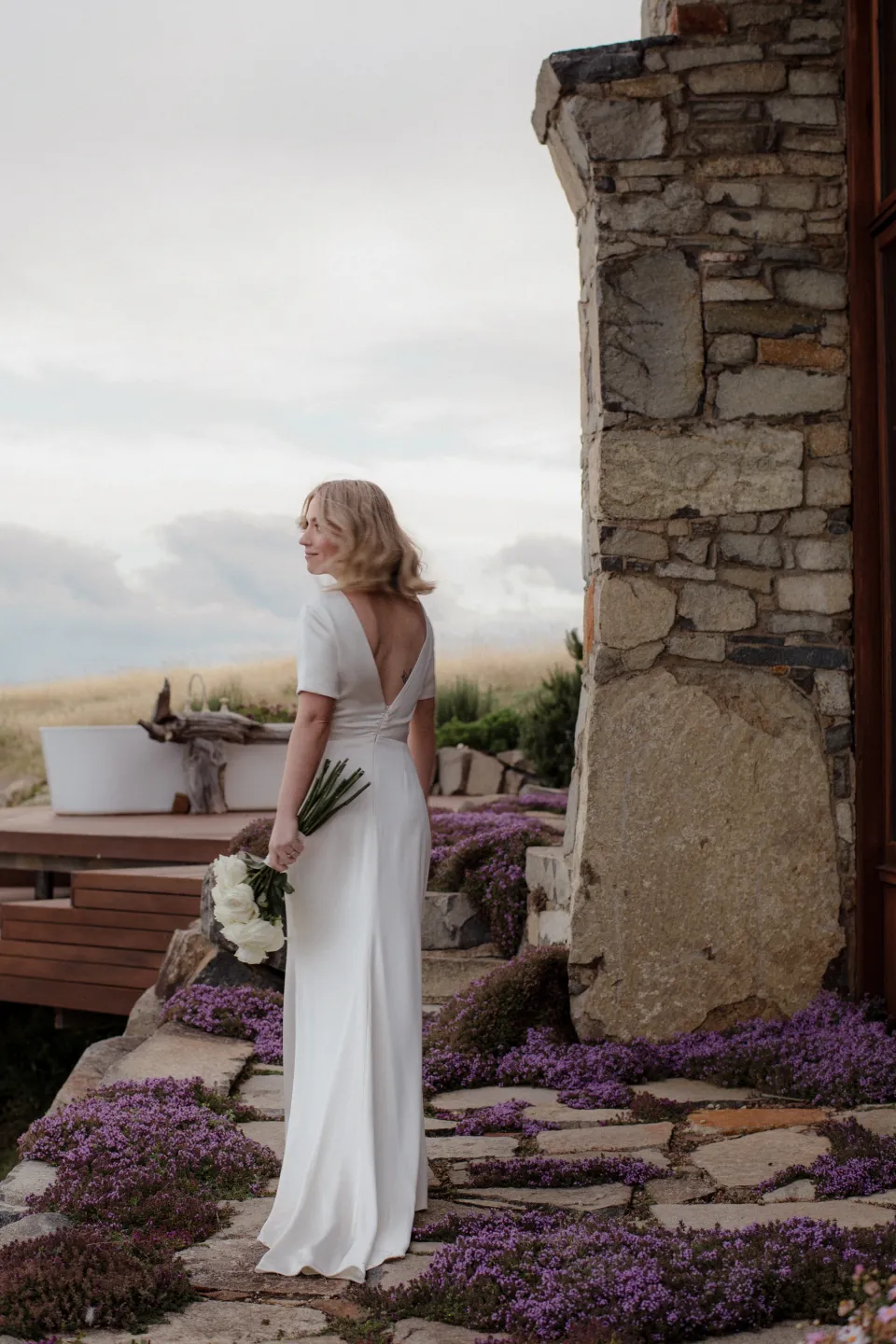 Jemmah turning towards the bay with her bouquet by The Romantics, at Thalia Haven on the East Coast of Tasmania, Australia, for an Elopement Collective elopement. Photograph by Joey Willis.