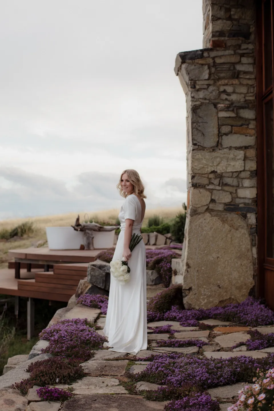 Jemmah smiling on the stone terrace above the bay, at Thalia Haven on the East Coast of Tasmania, Australia, for an Elopement Collective elopement. Photograph by Joey Willis.