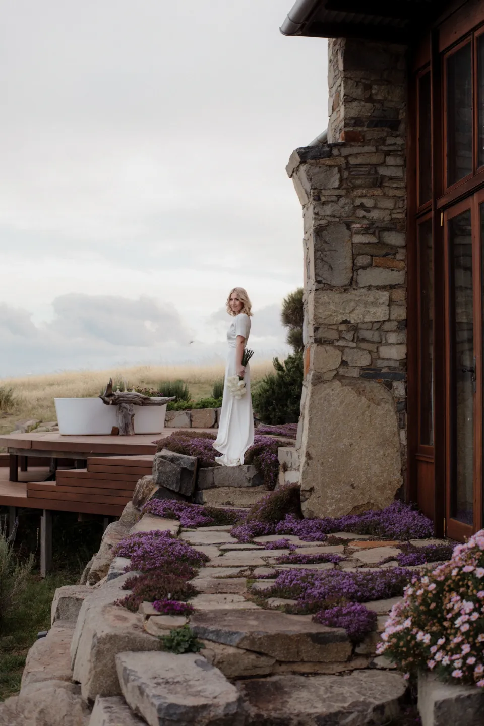 A wider view of Jemmah standing beside the stone building and outdoor bath, at Thalia Haven on the East Coast of Tasmania, Australia, for an Elopement Collective elopement. Photograph by Joey Willis.