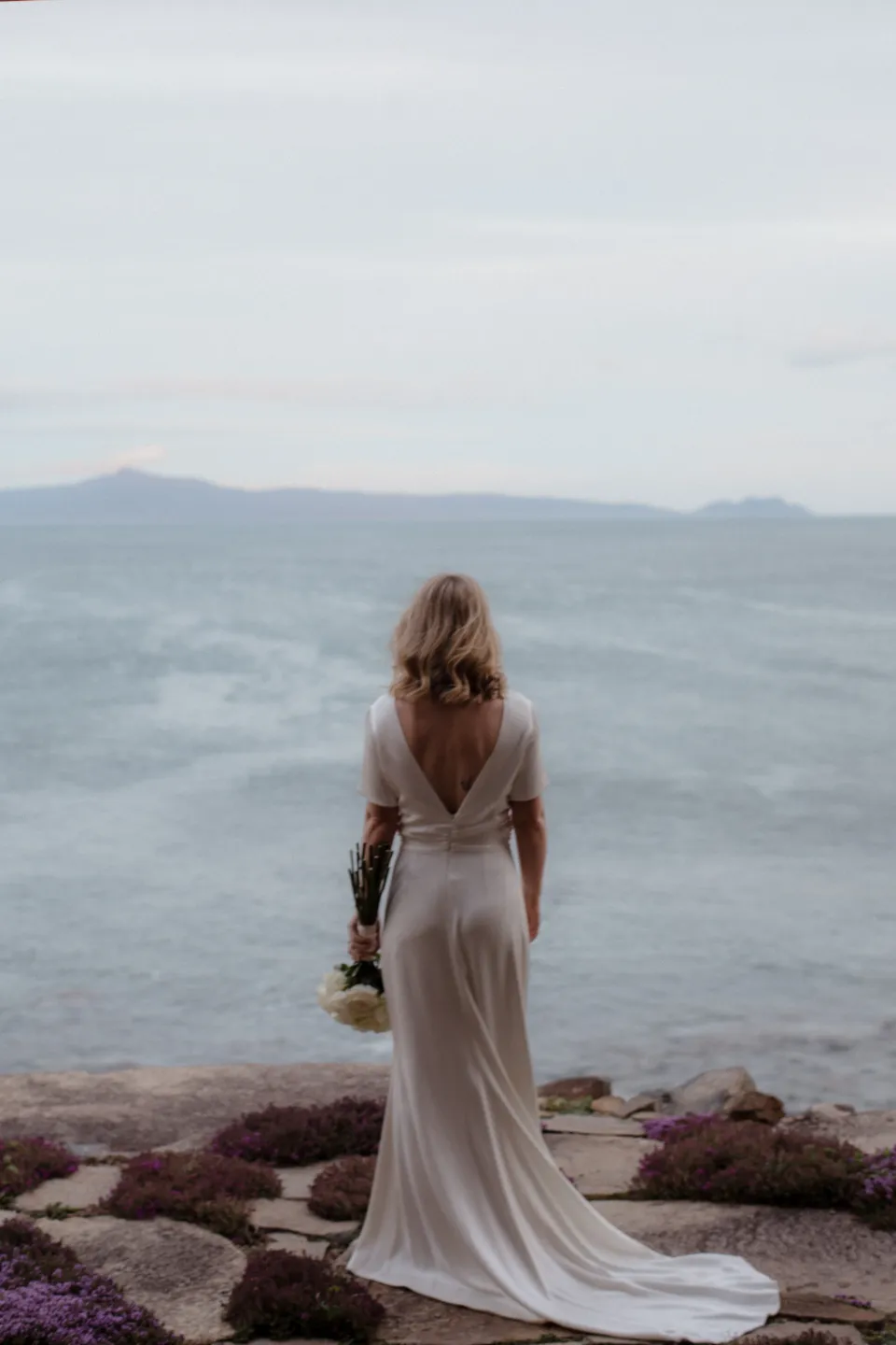 Jemmah facing Great Oyster Bay with her dress train spread across stone paving, at Thalia Haven on the East Coast of Tasmania, Australia, for an Elopement Collective elopement. Photograph by Joey Willis.