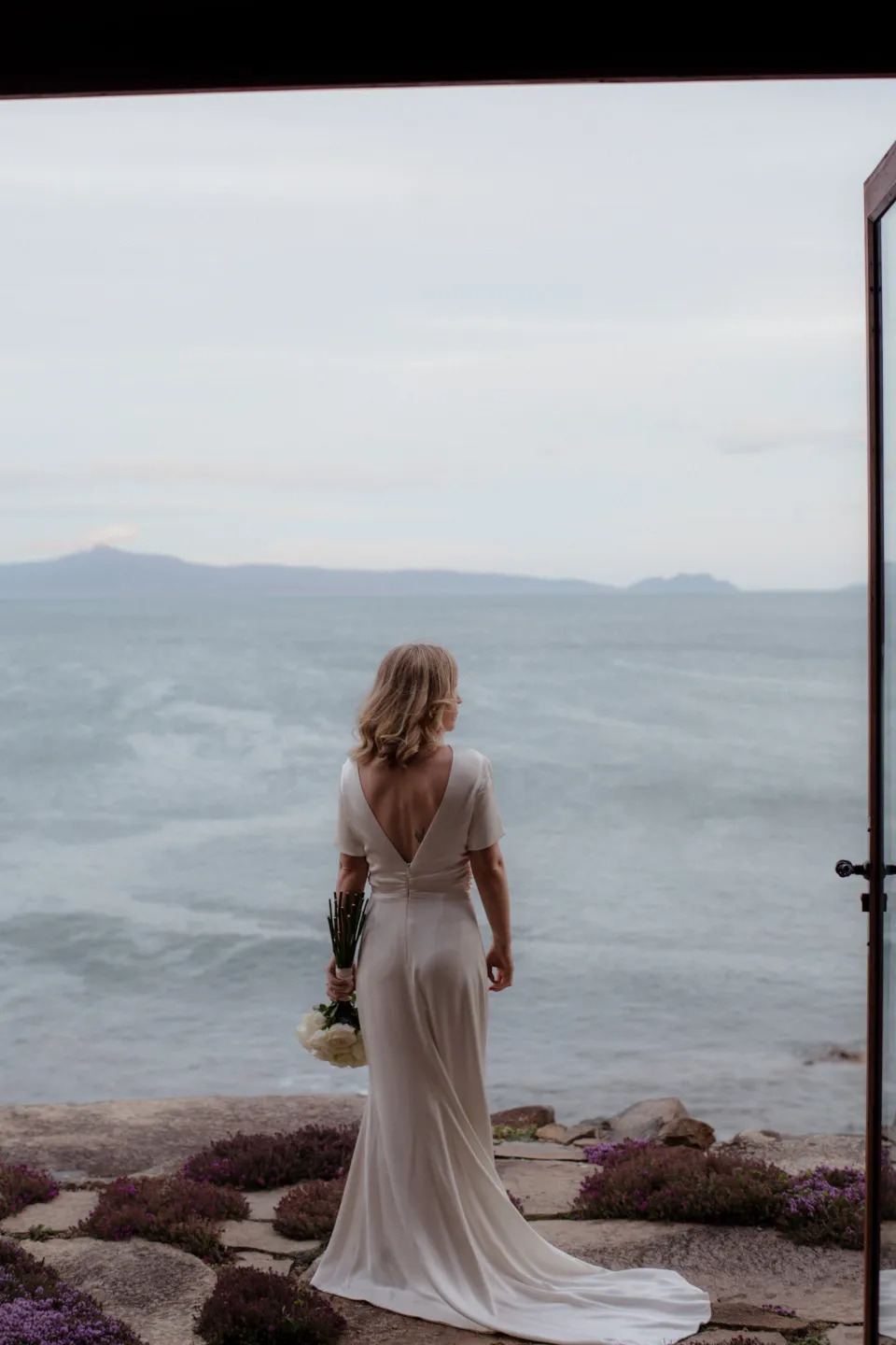 Jemmah framed by a doorway as she looks out to sea, at Thalia Haven on the East Coast of Tasmania, Australia, for an Elopement Collective elopement. Photograph by Joey Willis.