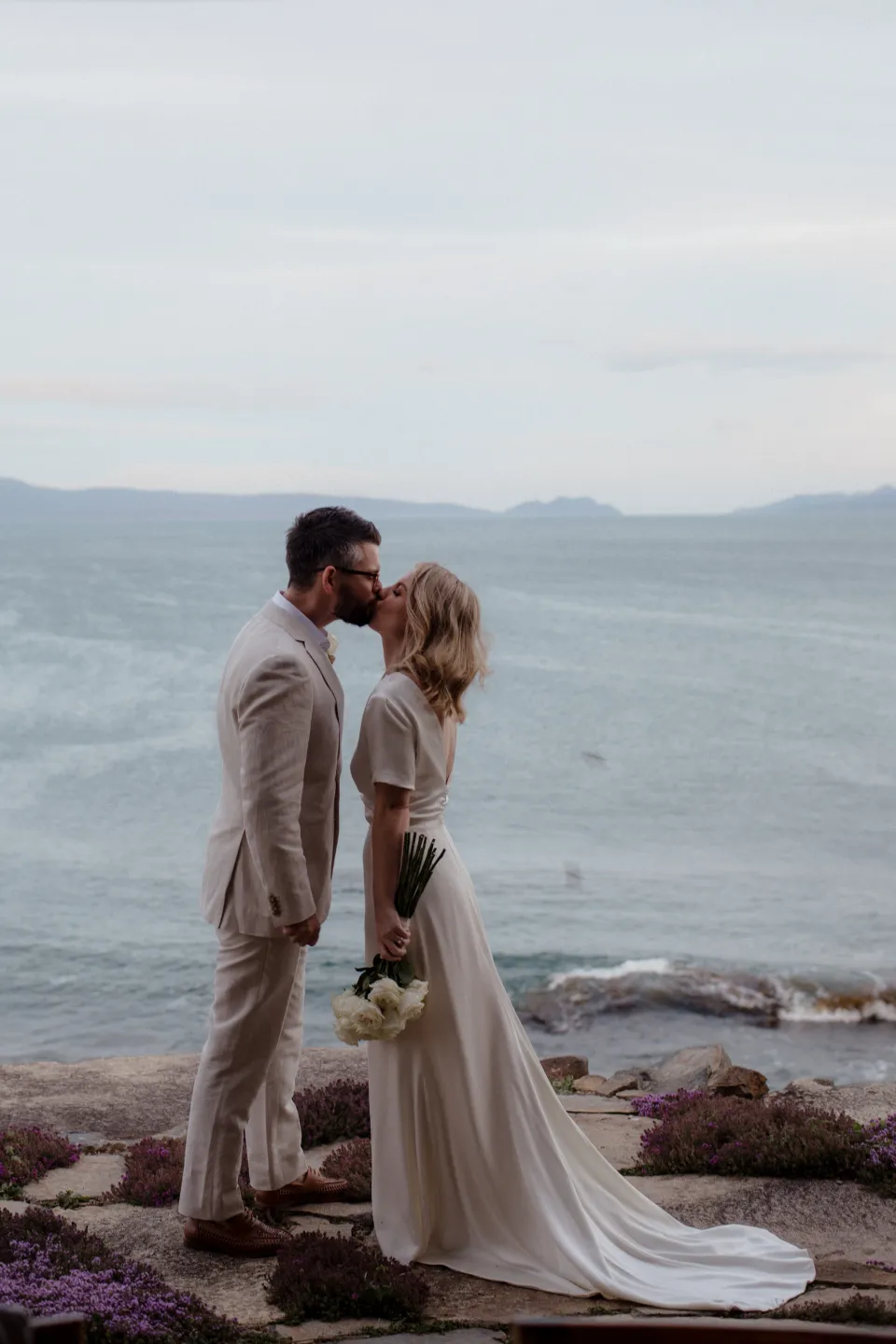 Jemmah and Jack kissing on the stone terrace beside the bay, at Thalia Haven on the East Coast of Tasmania, Australia, for an Elopement Collective elopement. Photograph by Joey Willis.