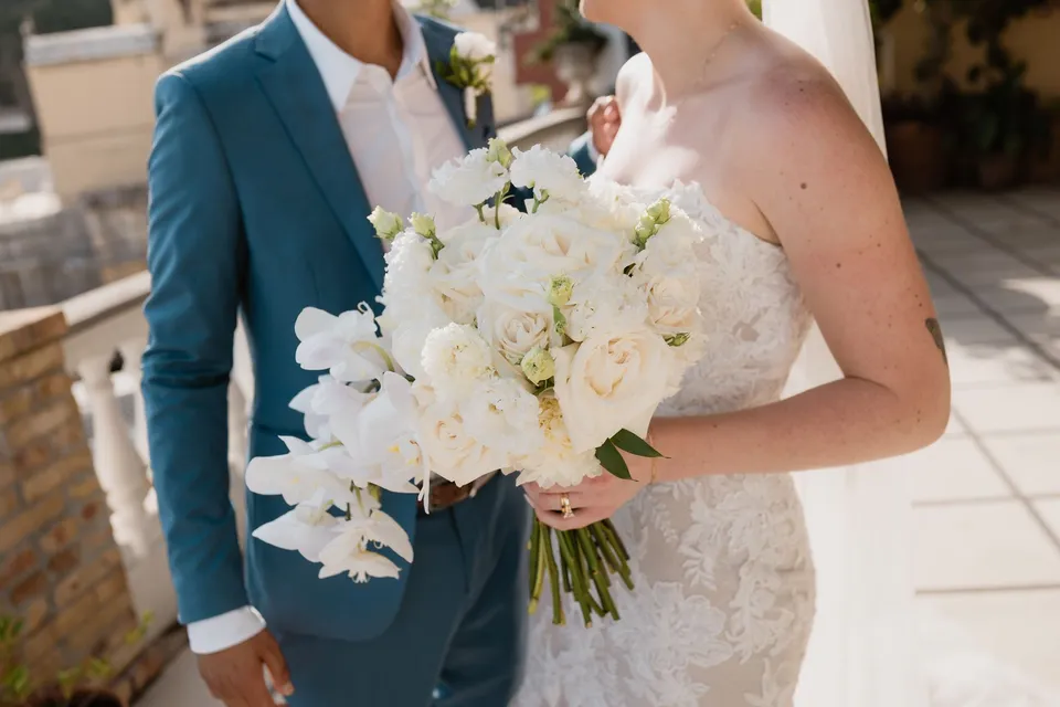 Close view of Skyler holding her white rose and cascading orchid bouquet by Flora Garden - Positano, beside Zach’s blue suit and white buttonhole, at Hotel Buca di Bacco in Positano, Amalfi Coast, Italy, for an Elopement Collective elopement. Photograph by Jason Corroto.