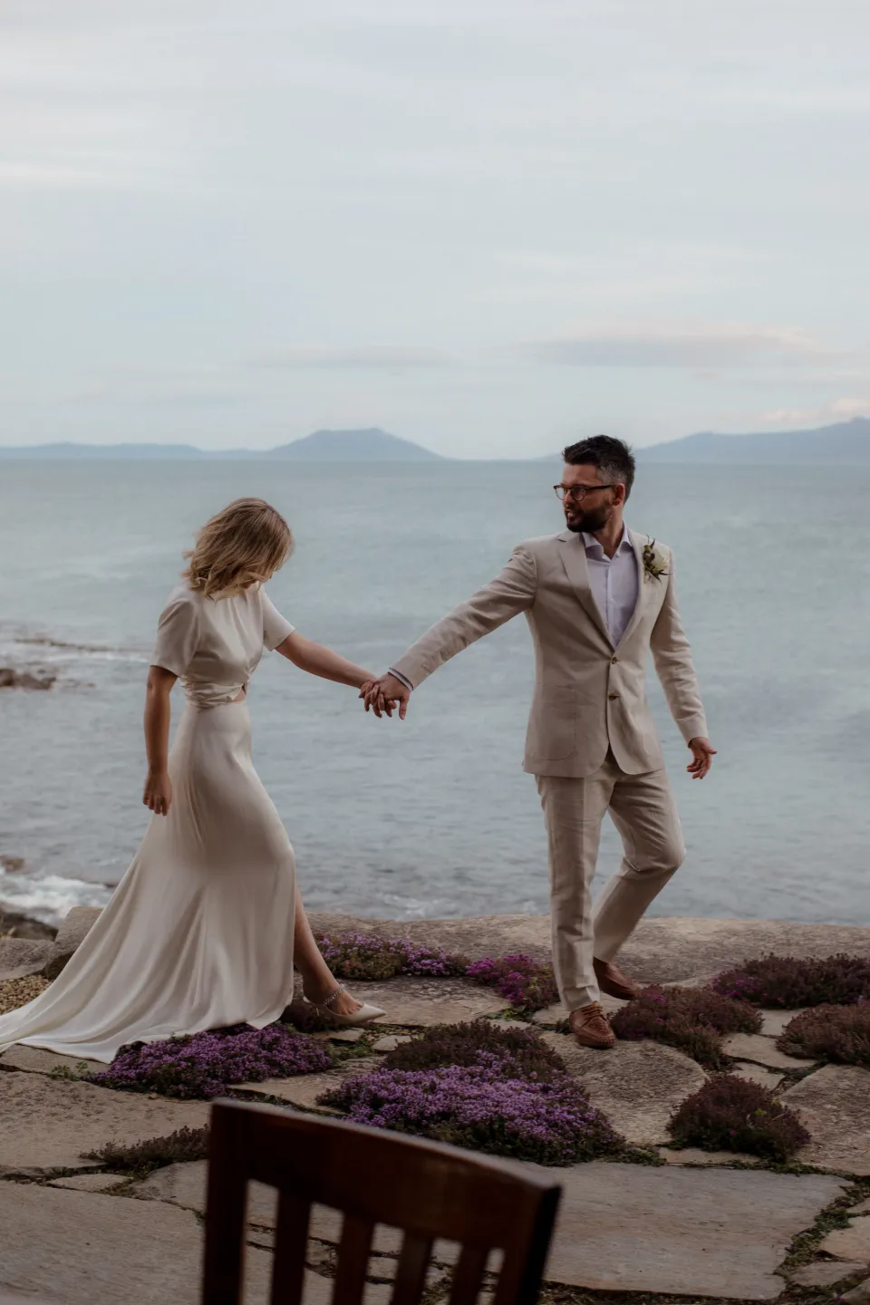 Jack taking Jemmah’s hand as they walk across the stone terrace, at Thalia Haven on the East Coast of Tasmania, Australia, for an Elopement Collective elopement. Photograph by Joey Willis.