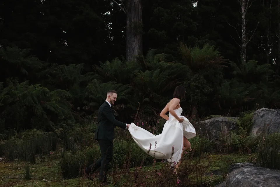 Ryan carrying Meg’s wedding dress train as she steps over a rock at the edge of the forest. At The Keep on Tasmania’s east coast, for an Elopement Collective elopement. Photograph by Jenna Agius.