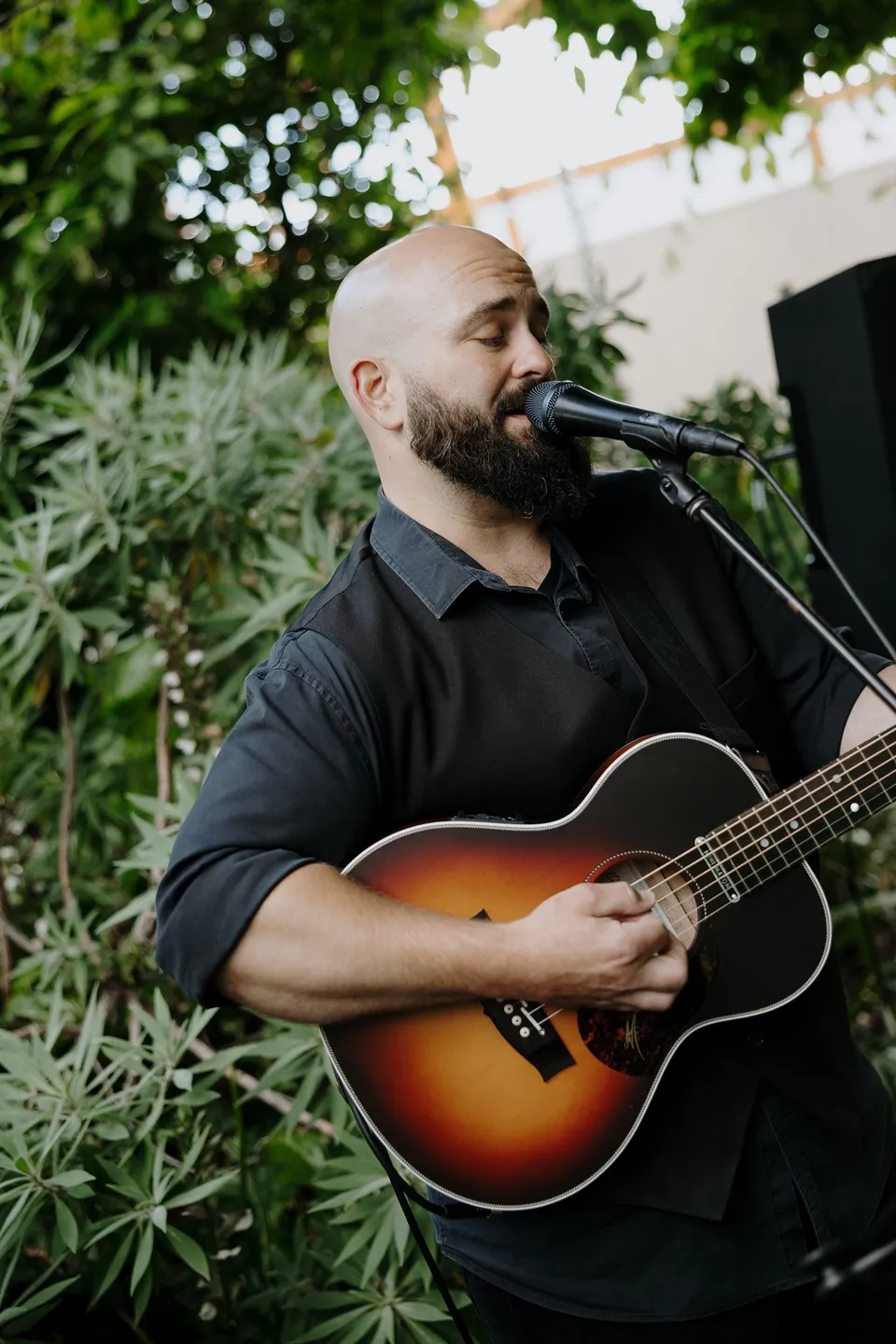 A musician singing into a microphone and playing an acoustic guitar in the hotel garden, at Islington Hotel in South Hobart, Tasmania, Australia, for an Elopement Collective summer elopement. Photograph by George Bowden.