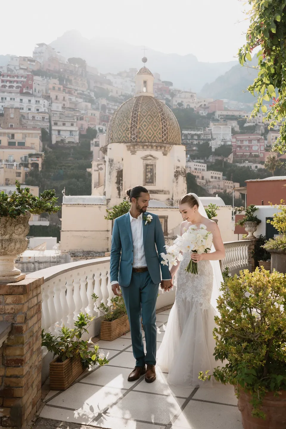 Skyler and Zach standing together on the terrace with her white bouquet by Flora Garden - Positano, beside the tiled church dome, at Hotel Buca di Bacco in Positano, Amalfi Coast, Italy, for an Elopement Collective elopement. Photograph by Jason Corroto.