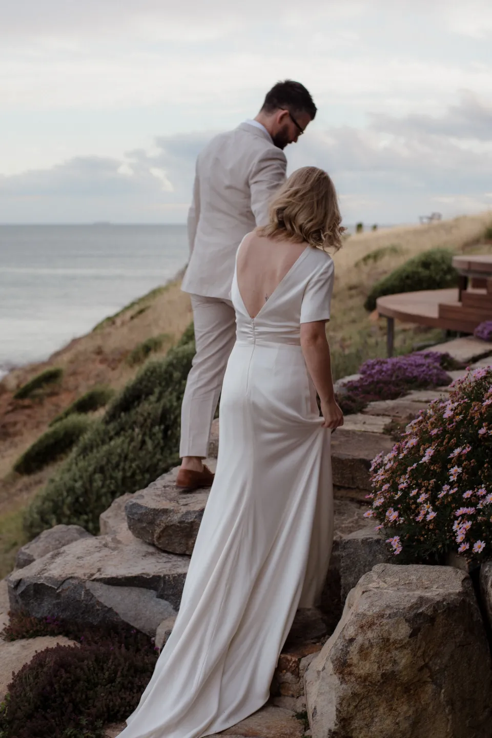 The couple walking up stone steps among garden flowers beside the house, at Thalia Haven on the East Coast of Tasmania, Australia, for an Elopement Collective elopement. Photograph by Joey Willis.