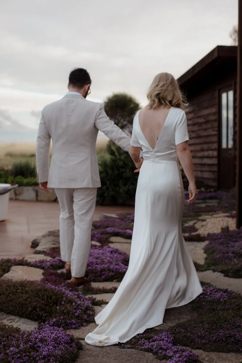 Rear view of Jemmah and Jack walking hand in hand beside the timber-clad building, at Thalia Haven on the East Coast of Tasmania, Australia, for an Elopement Collective elopement. Photograph by Joey Willis.