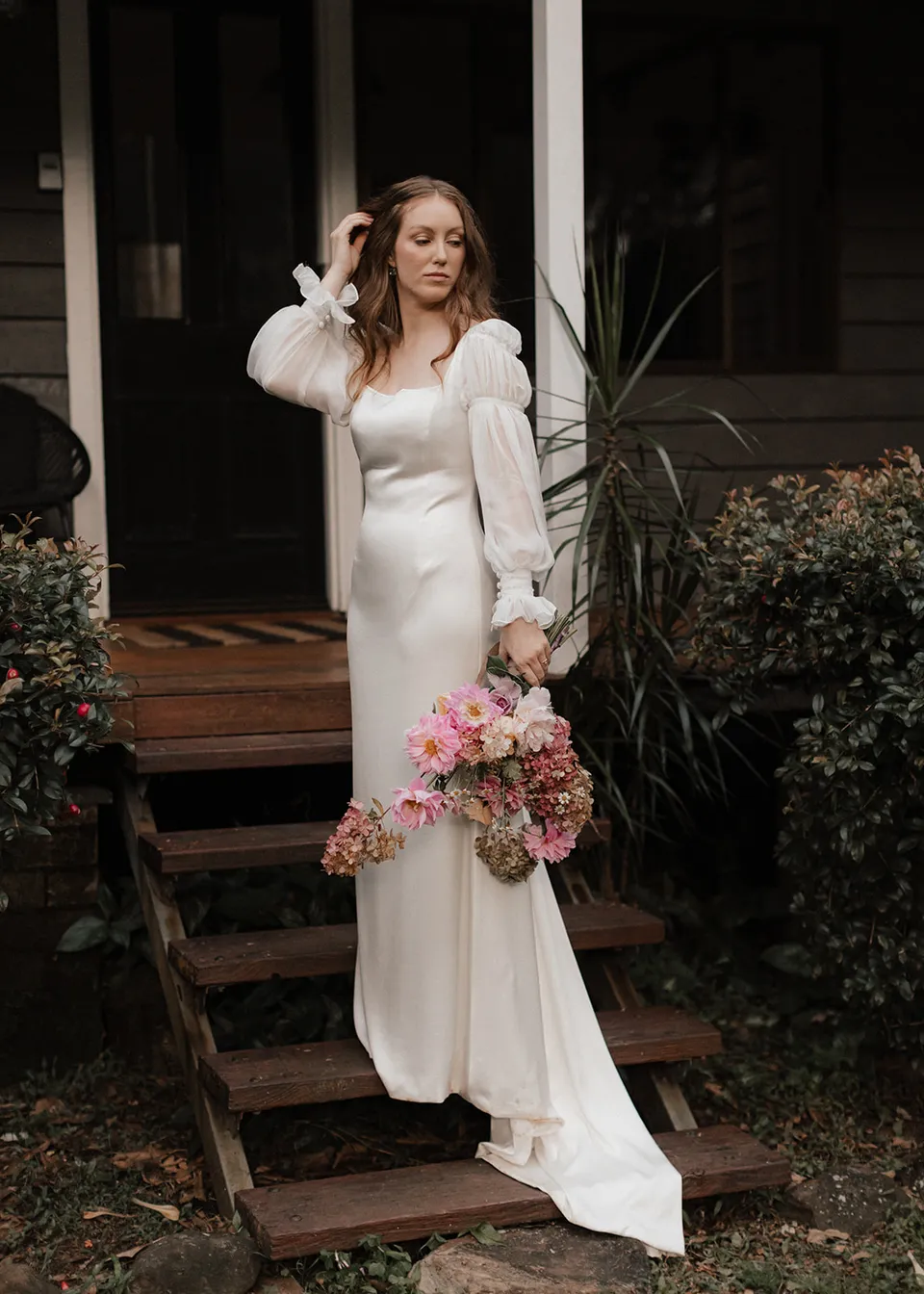 Elyse touching her hair while standing beside the timber steps. At Fig Tree Villa in Ewingsdale, Byron Bay, for an Elopement Collective elopement. Photograph by Bulb Creative.