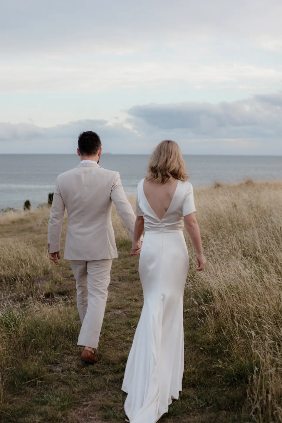 Jemmah and Jack walking towards the coastal meadow with the bay beyond, at Thalia Haven on the East Coast of Tasmania, Australia, for an Elopement Collective elopement. Photograph by Joey Willis.