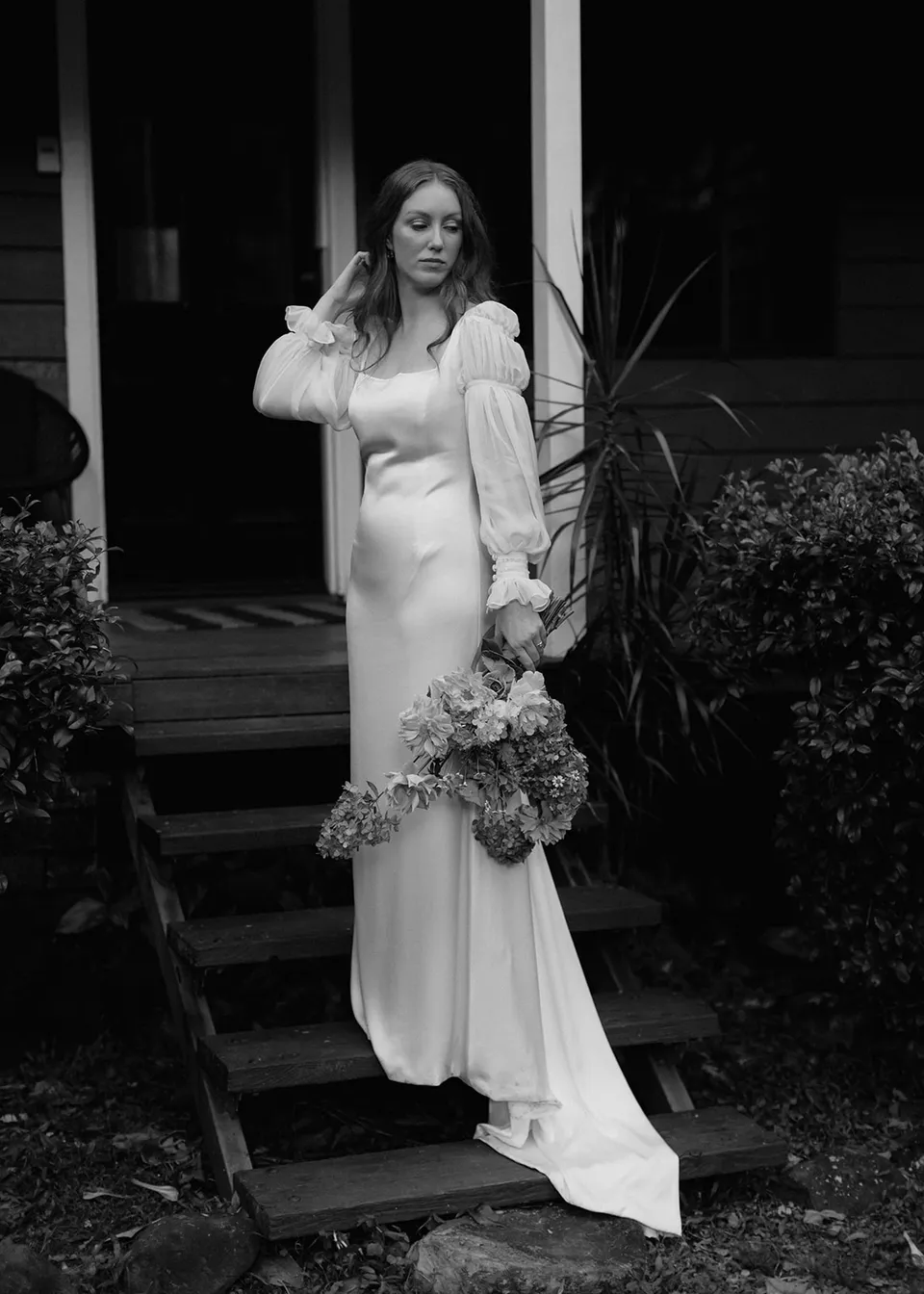 Black-and-white portrait of Elyse lifting her hand to her hair beside the verandah. At Fig Tree Villa in Ewingsdale, Byron Bay, for an Elopement Collective elopement. Photograph by Bulb Creative.