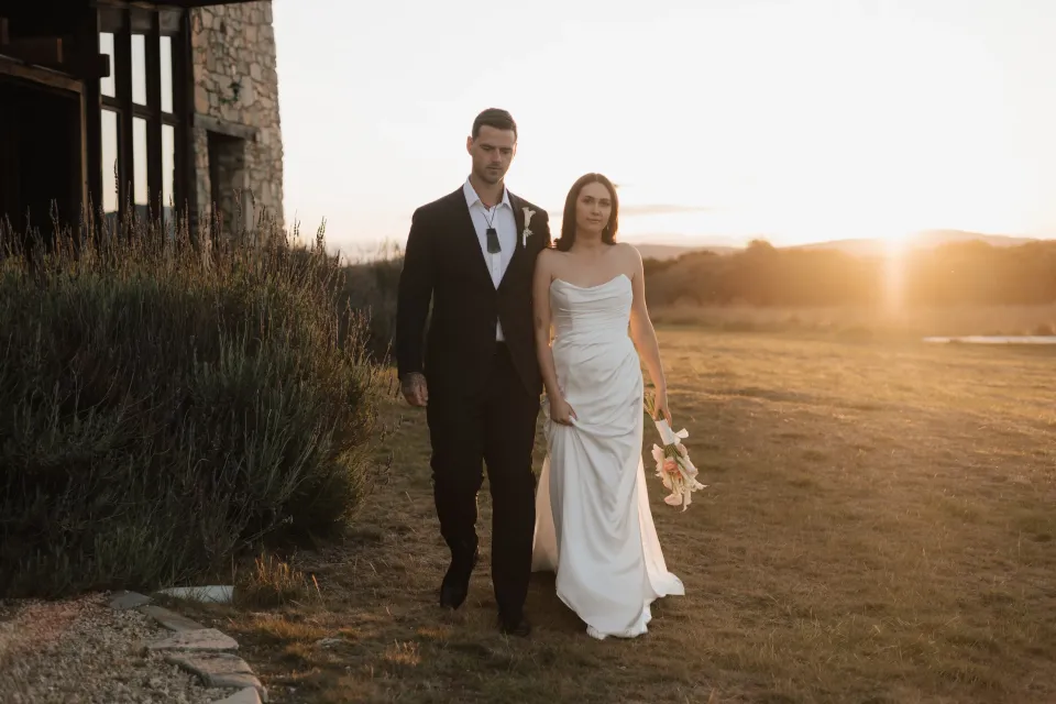 Dani and Anthony beside Thalia Haven’s stone house at sunset, with her bouquet by The Romantics and warm light across the grass, at Thalia Haven on Tasmania’s east coast, for an Elopement Collective elopement. Photograph by Bulb Creative.