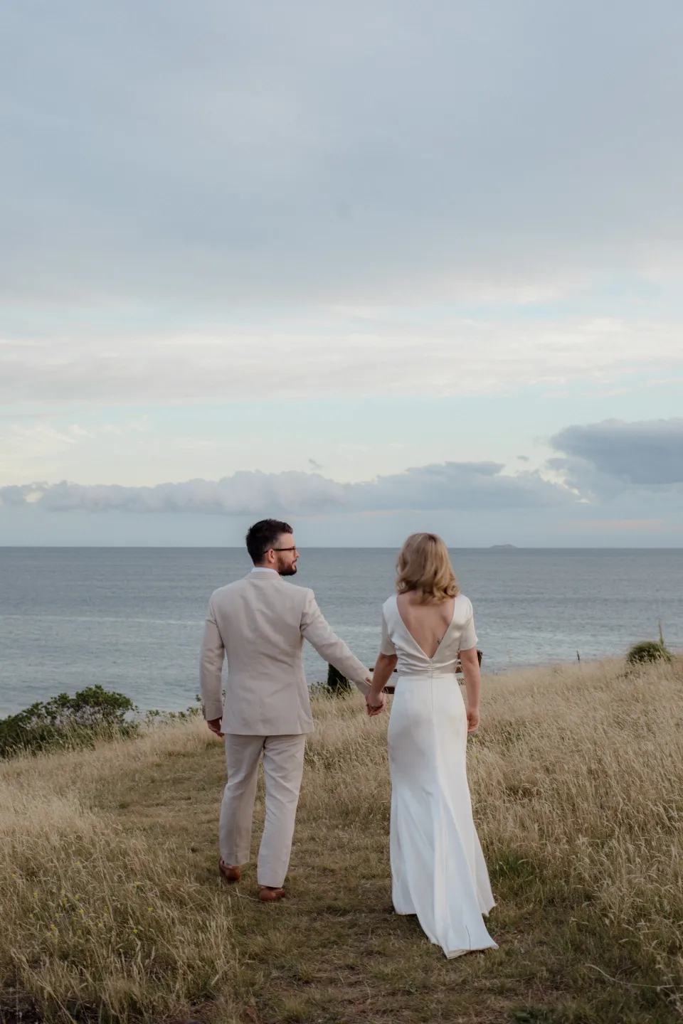 The couple standing together among golden grass with the sea behind them, at Thalia Haven on the East Coast of Tasmania, Australia, for an Elopement Collective elopement. Photograph by Joey Willis.