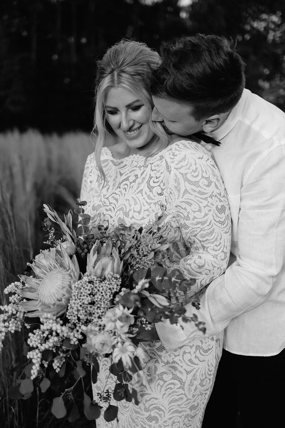 A black and white portrait of Matt kissing Laura’s cheek as she smiles and holds her Fifth Avenue Florist bouquet, in the Tweed Valley, NSW, for an Elopement Collective elopement. Photo by Bulb Creative.