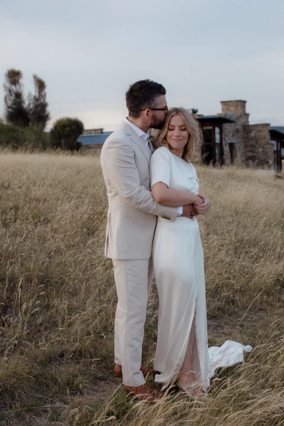 The couple embracing among tall grass with the stone house behind them, at Thalia Haven on the East Coast of Tasmania, Australia, for an Elopement Collective elopement. Photograph by Joey Willis.