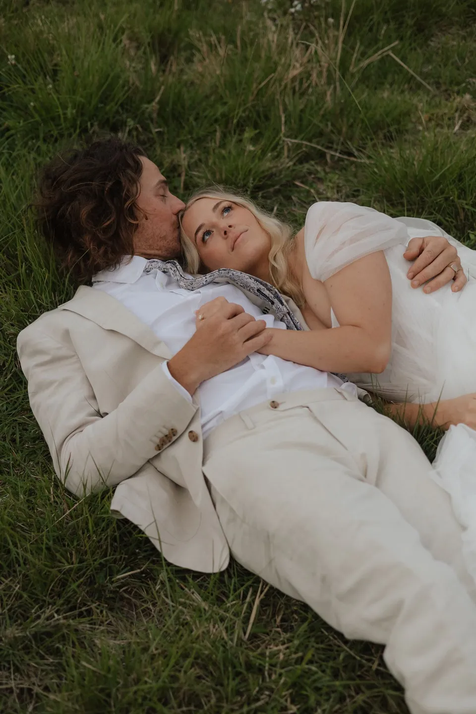 Bridget and James lying close together in the grass, smiling at each other. At Cudgera Creek in northern New South Wales, for an Elopement Collective elopement. Photograph by Bulb Creative.