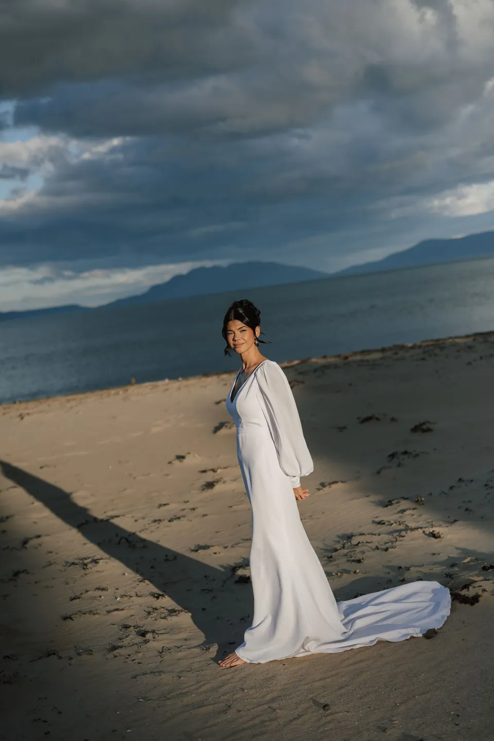 Renee standing barefoot on the beach, with her wedding dress trailing across the sand and dark clouds over the distant hills. At Piermont Retreat on Tasmania’s east coast, for an Elopement Collective elopement. Photograph by Bulb Creative.
