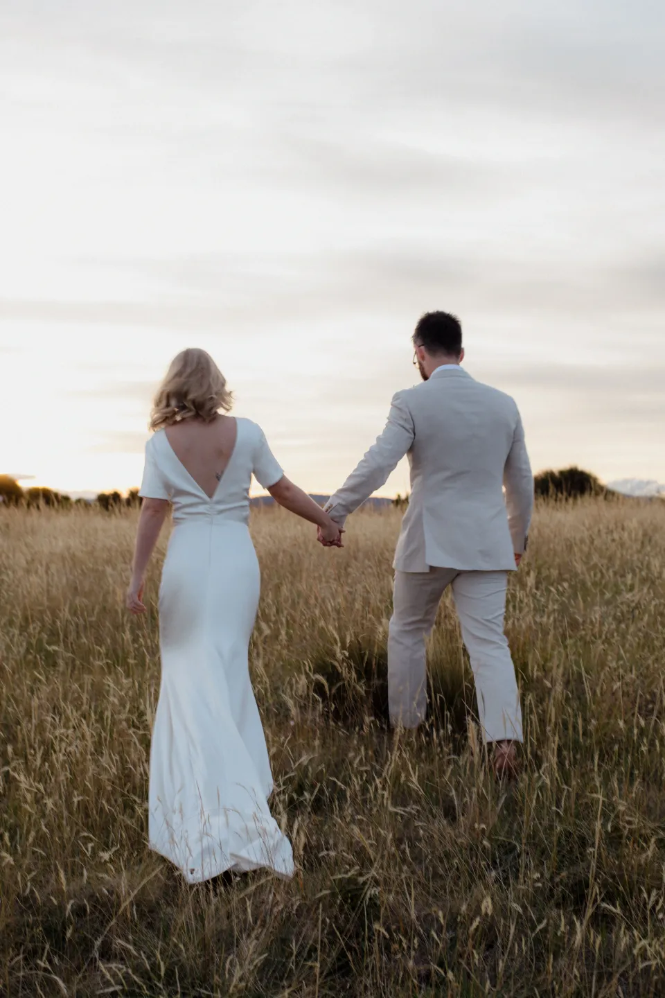 Jemmah and Jack walking away into the meadow as the light warms, at Thalia Haven on the East Coast of Tasmania, Australia, for an Elopement Collective elopement. Photograph by Joey Willis.